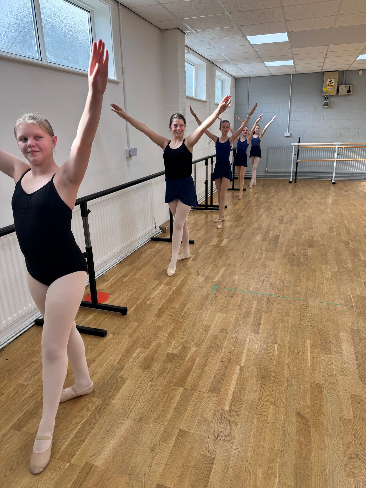 A line of ballet students at the barre with arms raised in the studio