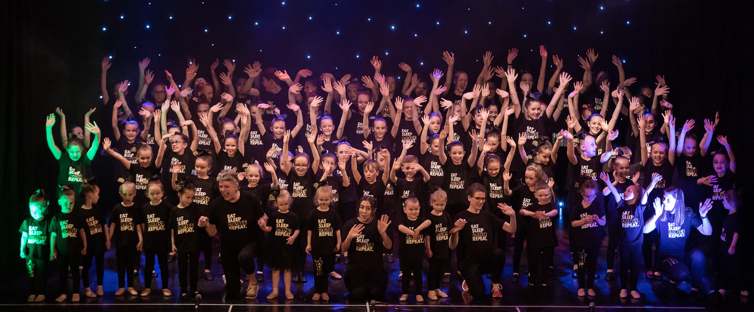 The whole school on stage under coloured lights, arms raised at the end of a McCaw show